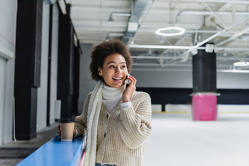 Positive african american woman talking on smartphone and holding paper cup on ice rink