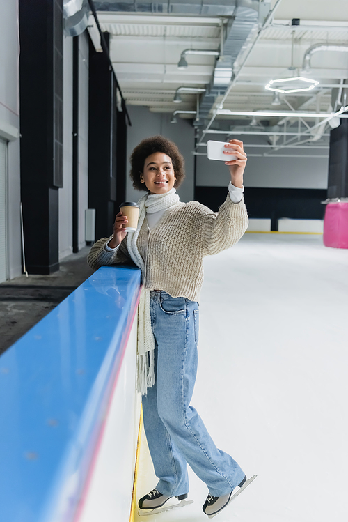 African american woman in ice skates holding coffee to go and taking selfie on smartphone on ice rink