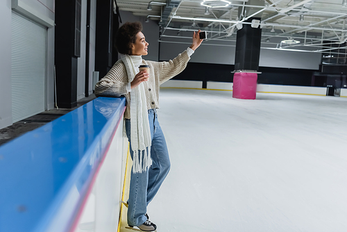 Side view of african american woman in ice skates holding paper cup and taking selfie on smartphone on ice rink