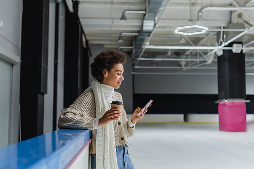 Side view of african american woman in warm scarf holding coffee to go and using cellphone on ice rink