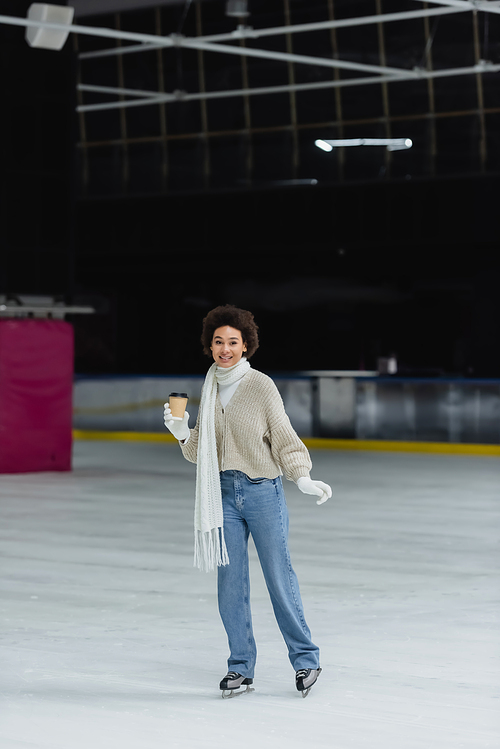 Smiling african american woman holding coffee to go while ice skating on rink