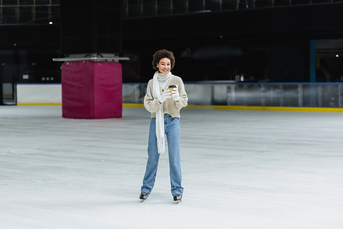 Cheerful african american woman holding paper cup and looking at camera on ice rink