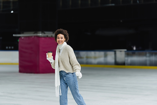 Smiling african american woman in scarf and gloves holding paper cup and looking at camera on ice rink