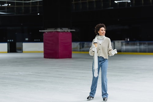 Young african american woman ice skating and holding coffee to go on rink