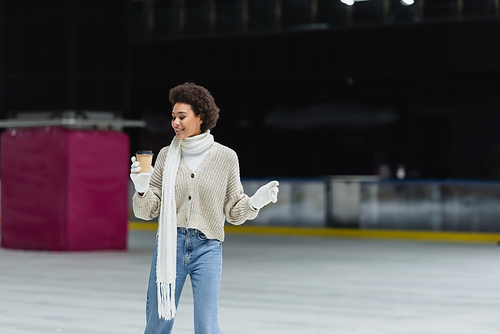 Positive african american woman in gloves and warm clothes holding coffee to go on ice rink