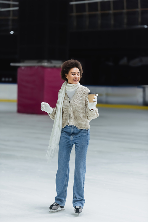 Smiling african american woman ice skating and looking at takeaway coffee in paper cup on ice rink