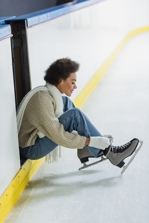 Side view of positive african american woman tying shoelaces of ice skates on rink