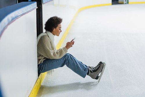 Side view of african american woman in ice skates using smartphone on rink