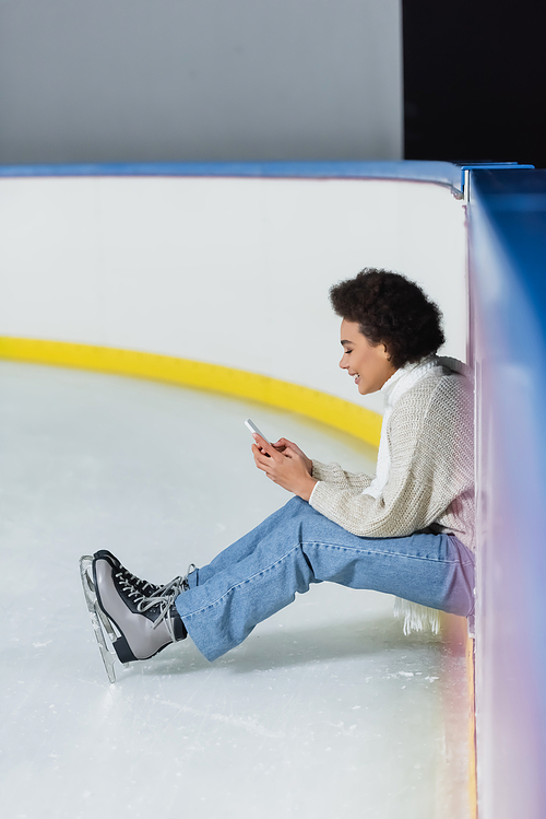 Side view of cheerful african american woman in ice skates and scarf using smartphone on rink