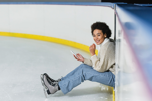 Positive african american woman holding smartphone and looking at camera on ice rink