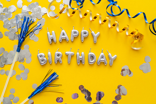 Top view of candles in shape of Happy Birthday lettering near confetti and serpentine on yellow background