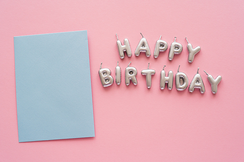 Top view of greeting card near candles in shape of Happy Birthday lettering on pink background