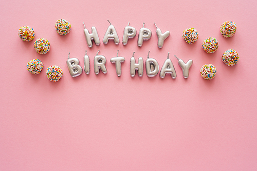 Top view of candles in shape of Happy Birthday lettering near colorful sweets on pink background