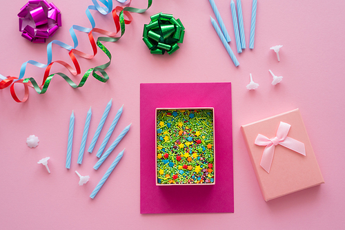 Top view of sprinkles in gift box near candles and serpentine on pink background