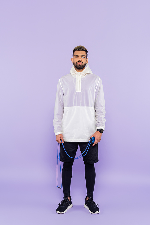 full length of young african american man in trendy sportswear holding skipping rope on purple