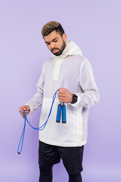 young african american man in trendy sportswear holding skipping rope isolated on purple