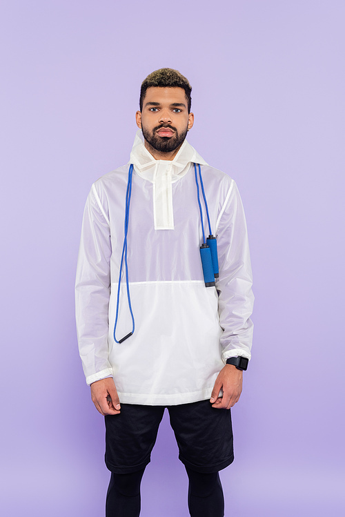 young african american man in stylish sportswear standing with skipping rope isolated on purple