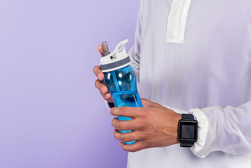 partial view of african american man in trendy sportswear holding sports bottle isolated on purple