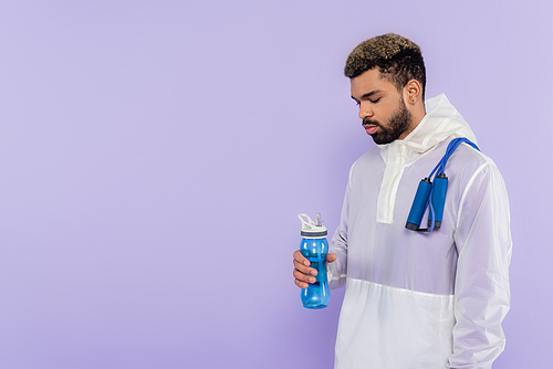 african american man in sportswear with skipping rope holding sports bottle isolated on purple