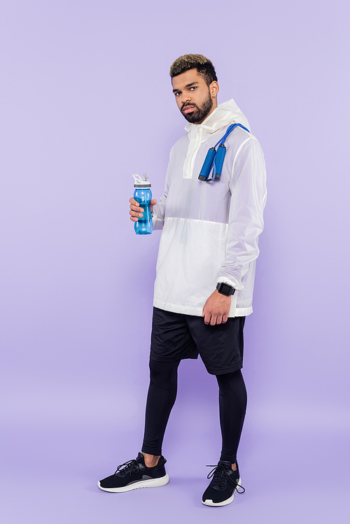 full length of african american man in sportswear standing with jumping rope and holding sports bottle on purple