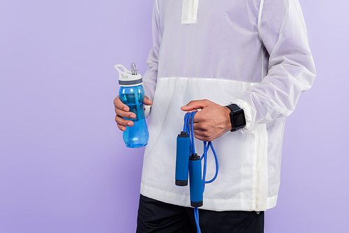 cropped view of african american man in trendy sportswear holding sports bottle and jumping rope isolated on purple