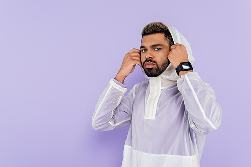 african american sportsman in stylish sportswear wearing hood looking at camera isolated on purple