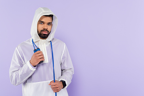 african american sportsman in stylish sportswear with hood looking at camera and holding skipping rope isolated on purple