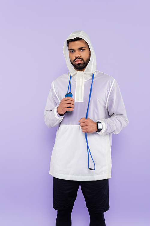 african american sportsman in sportswear with hood looking at camera and holding jumping rope isolated on purple