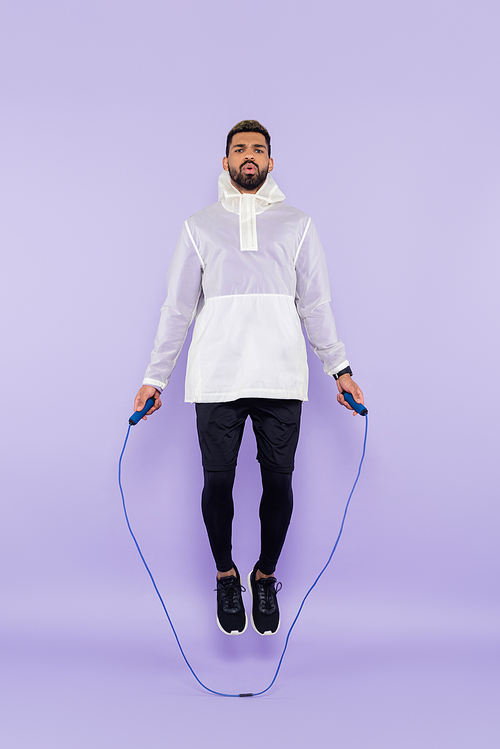 full length of african american sportsman in sportswear jumping with skipping rope on purple