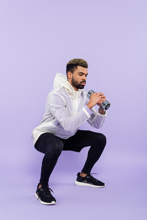 full length of bearded african american sportsman doing squat with dumbbell on purple