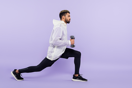 full length of bearded african american man doing lunges with dumbbells on purple