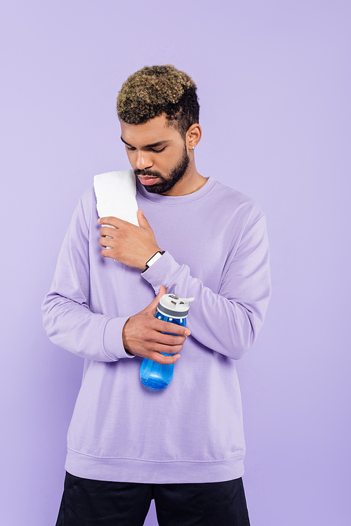 bearded african american man in sweater with white towel holding sport bottle with water isolated on purple