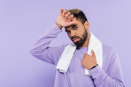 tired african american man in sweater holding white towel and wiping sweat with hand isolated on purple