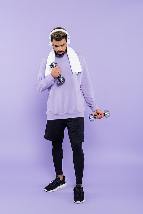 full length of bearded african american man in headphones exercising with dumbbells on purple