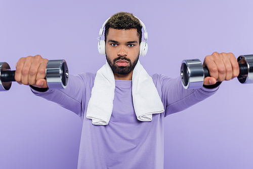 young african american man in headphones exercising with dumbbells isolated on purple