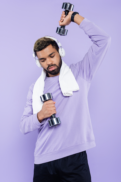 bearded african american man in wireless headphones exercising with dumbbells isolated on purple