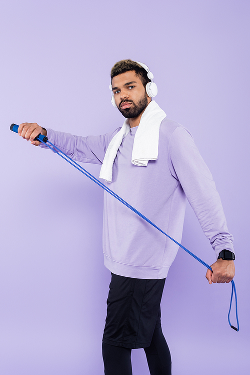 bearded african american man in wireless headphones holding jumping rope isolated on purple