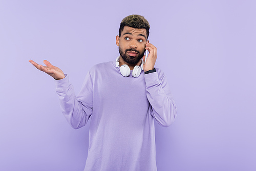 bearded african american man with wireless headphones talking on smartphone isolated on purple