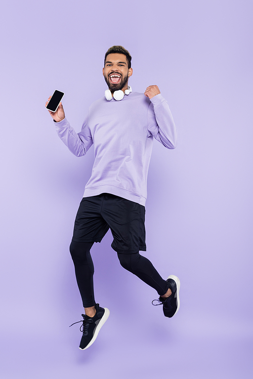 excited african american man with wireless headphones holding smartphone and jumping on purple