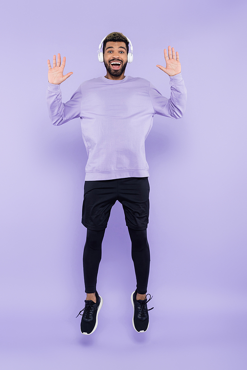 full length of excited african american man in wireless headphones jumping on purple