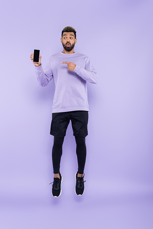 full length of surprised african american man pointing at smartphone and levitating on purple