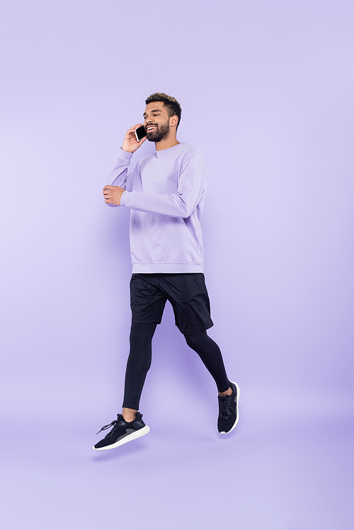 full length of happy african american man talking on smartphone and levitating on purple