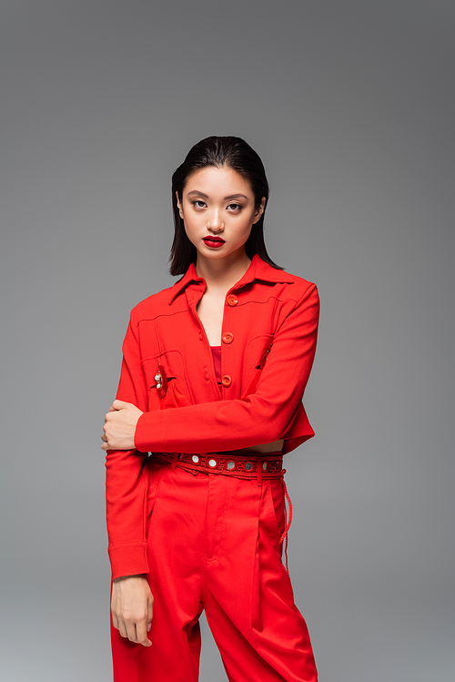 brunette asian woman in red trendy suit looking at camera isolated on grey