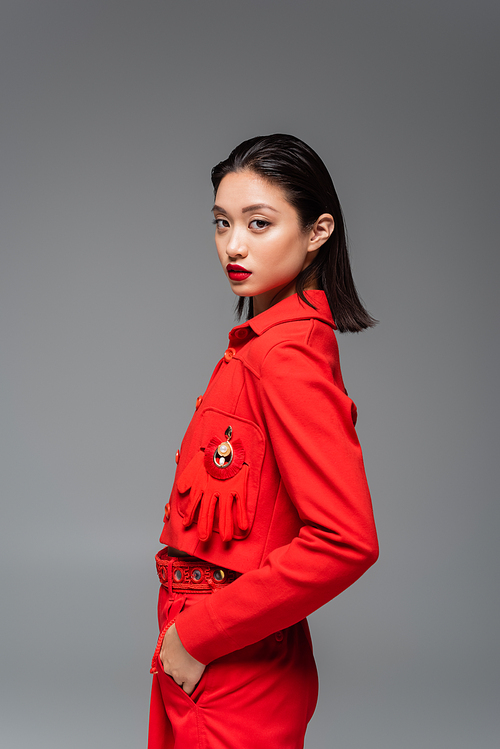 brunette asian model in red jacket decorated with glove posing with hand in pocket isolated on grey