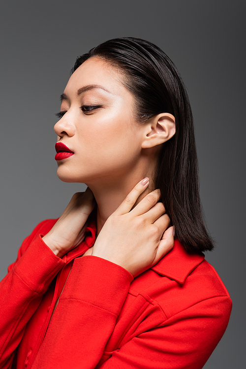 portait of asian woman in red jacket posing with hands on neck isolated on grey