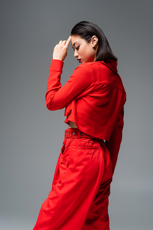 side view of asian woman in red stylish attire touching forehead isolated on grey