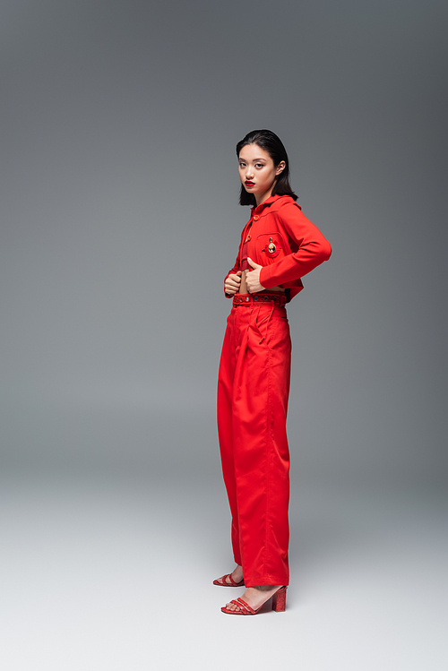 full length of young asian woman in red jacket and trousers looking at camera on grey background