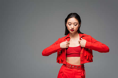 brunette asian woman touching red fashionable jacket isolated on grey