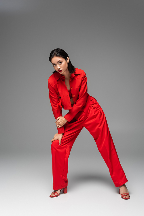 full length of stylish asian woman in red jacket and trousers looking at camera while posing on grey background