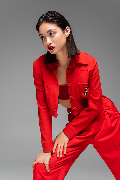 brunette asian woman in red elegant suit looking away while posing isolated on grey